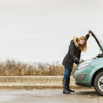 Girl standing over a broken down car looking under the hood.